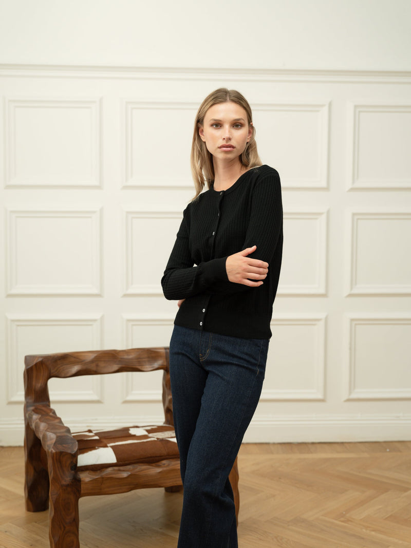 Woman wearing a Ninepine black merino wool cardigan with dark blue denim jeans against an off-white wainscoted wall