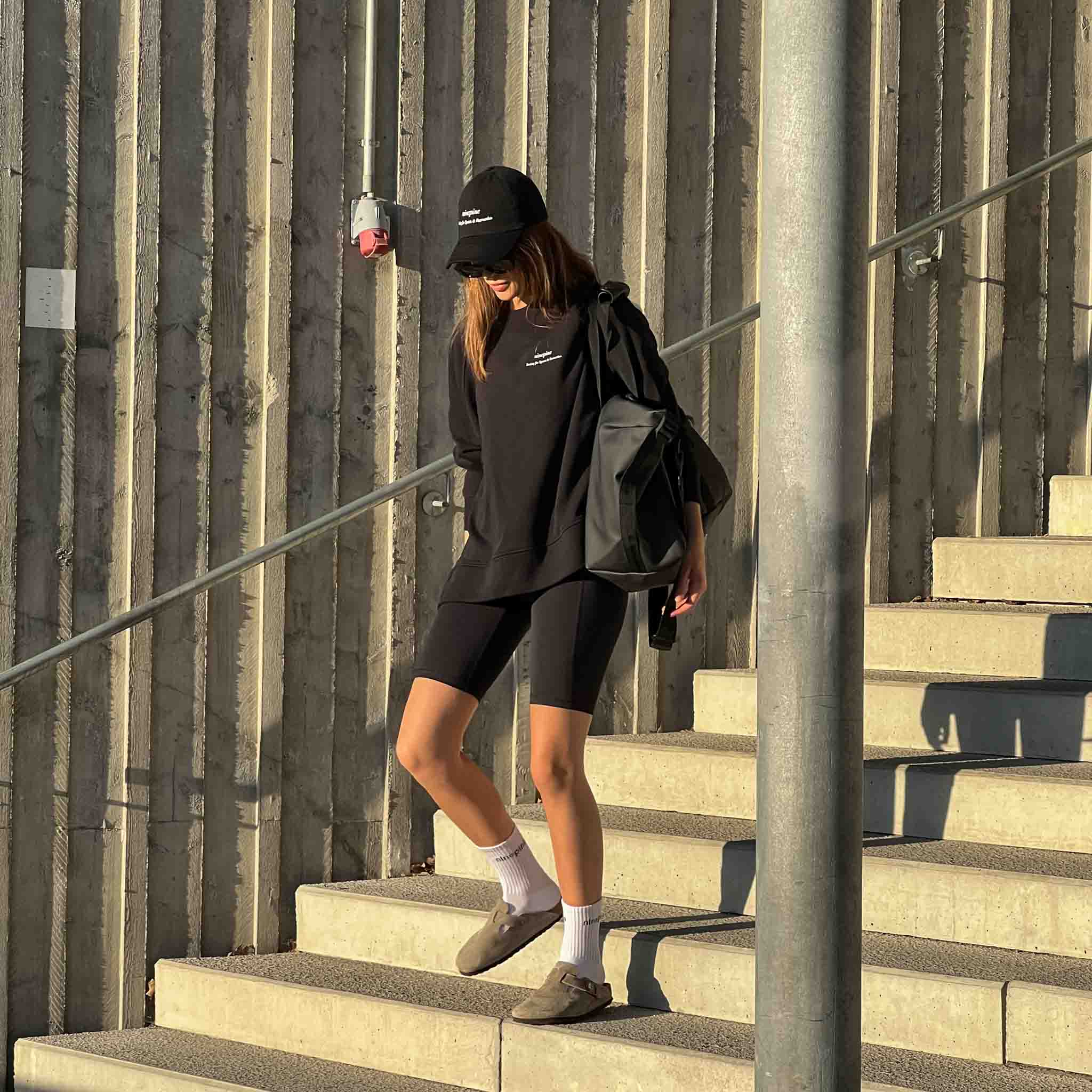 Woman in a black ninepine cap, sweater, biker shorts and sandles walking down concrete steps wiith sunglasses on and a gym bag on her left shoulder