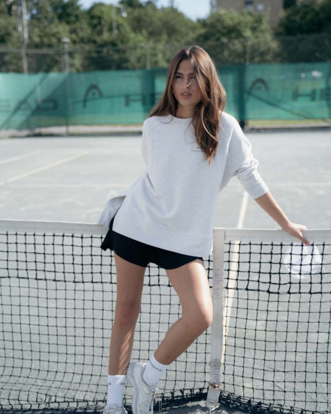Woman posing on tennis court net in shorts and oversized sweater