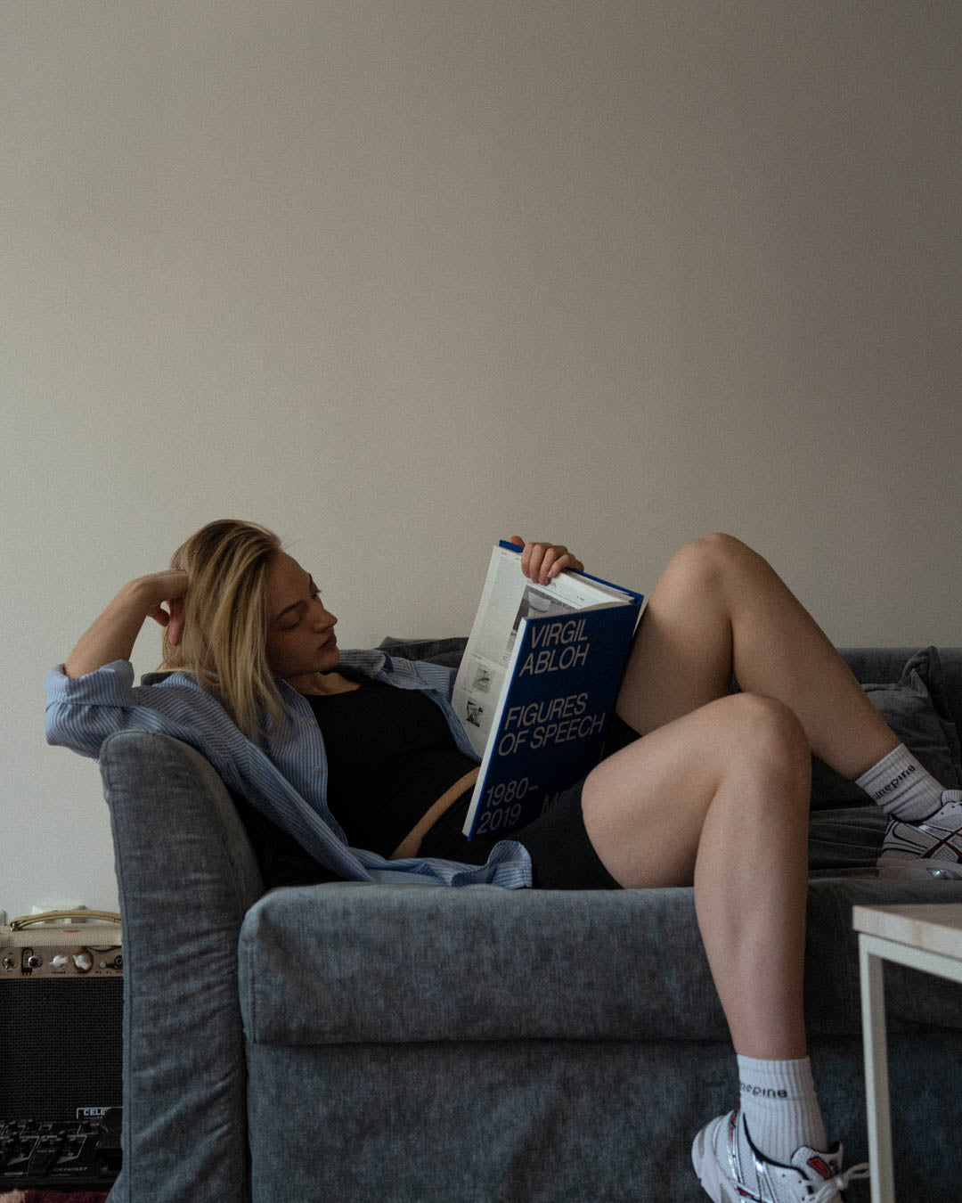 Blonde woman in biker shorts reading a book on a blue couch