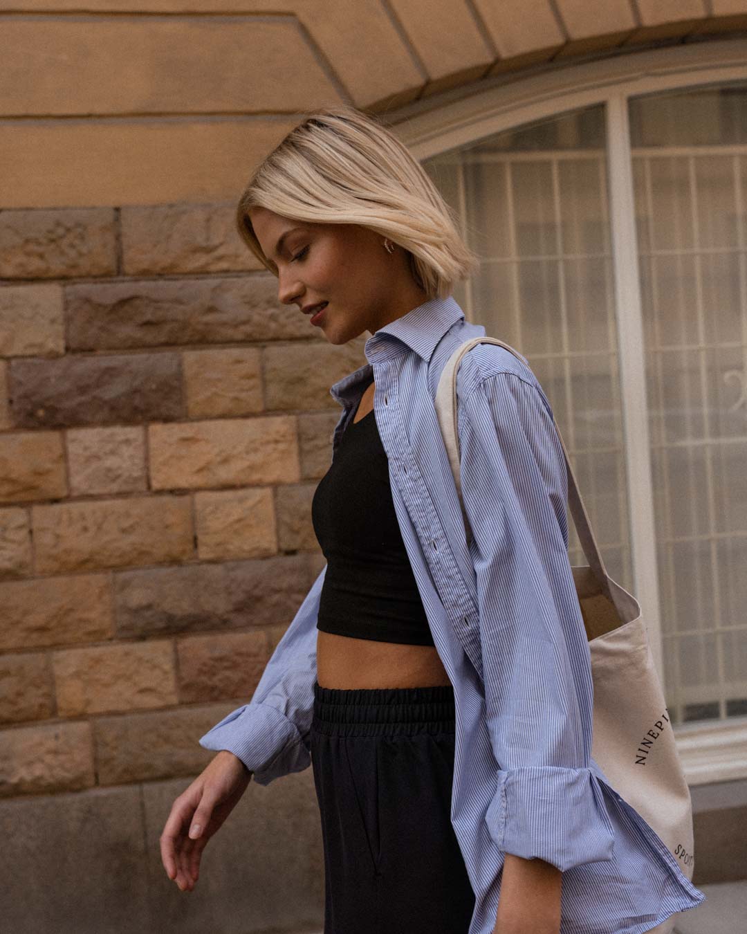 profile close up of a blonde woman in a blue dress shirt and ninepine crop tank walking down a street