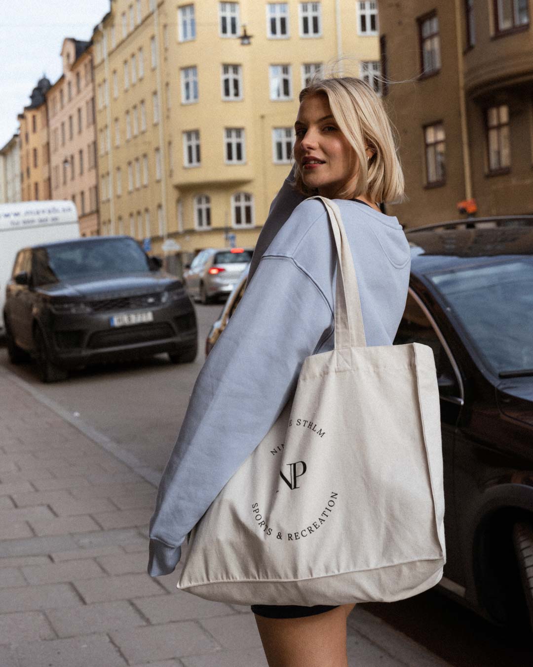 Blonde woman in biker shorts, blue sweater and a tote bag walking down a footpath in Stockholm