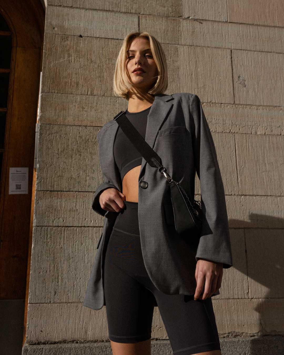 Blonde woman in biker shorts, suit jacket and handbag posing infront of a building
