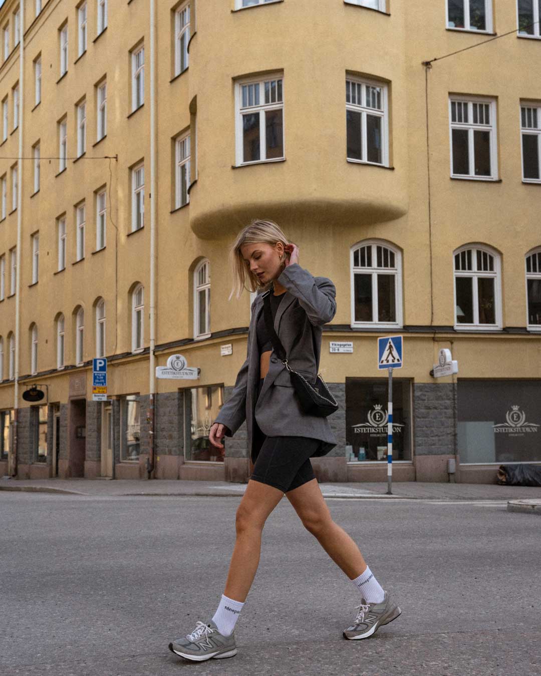Blonde woman in biker shorts, suit jacket and handbag walking across a Stockholm street
