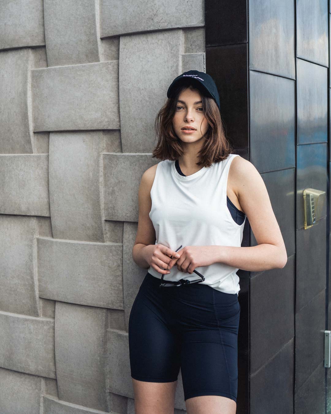 Woman in a black cap, white tank top and biker shorts leaning on a wall
