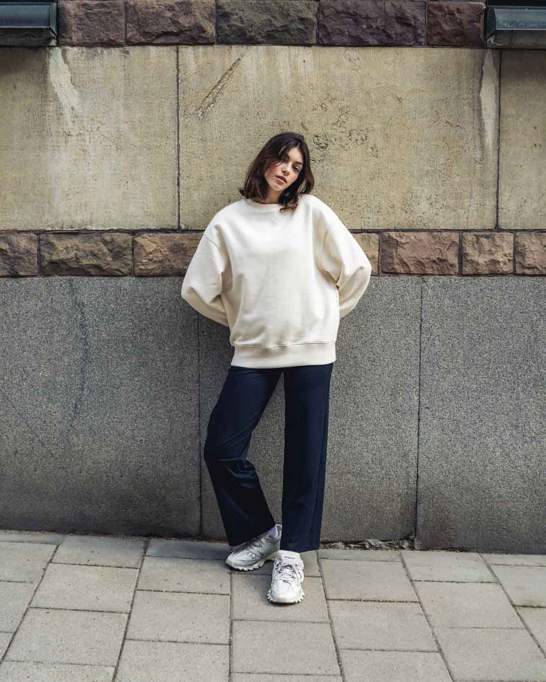 Woman in straight pants and over-sized cream sweatshirt leaning against a stone wall