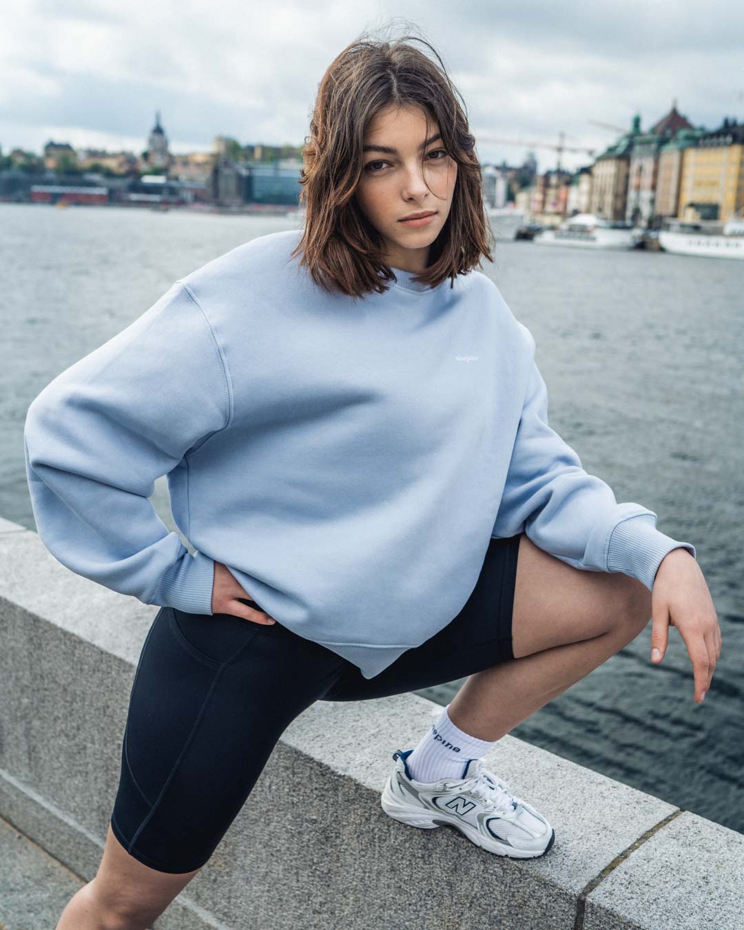Woman in a blue ninepine sweatshirt and biker shorts by the waters edge in Stockholm