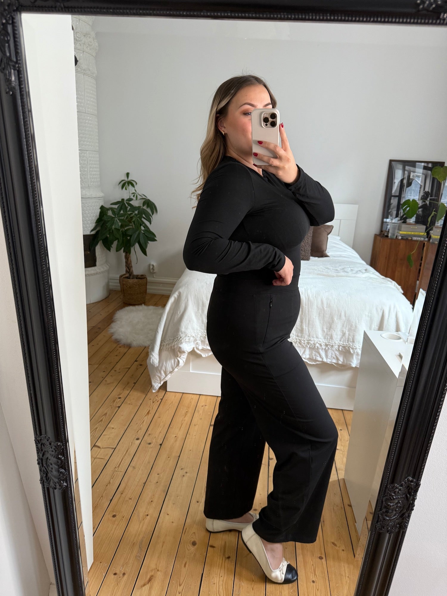 Eine kurvige Frau macht ein Spiegel-Selfie in einer schwarzen „Ninepine Asana Relaxed“-Hose und einem passenden langärmeligen, weichen Oberteil, kombiniert mit zweifarbigen Ballerinas – ein schlichtes, modernes Outfit für den Alltag