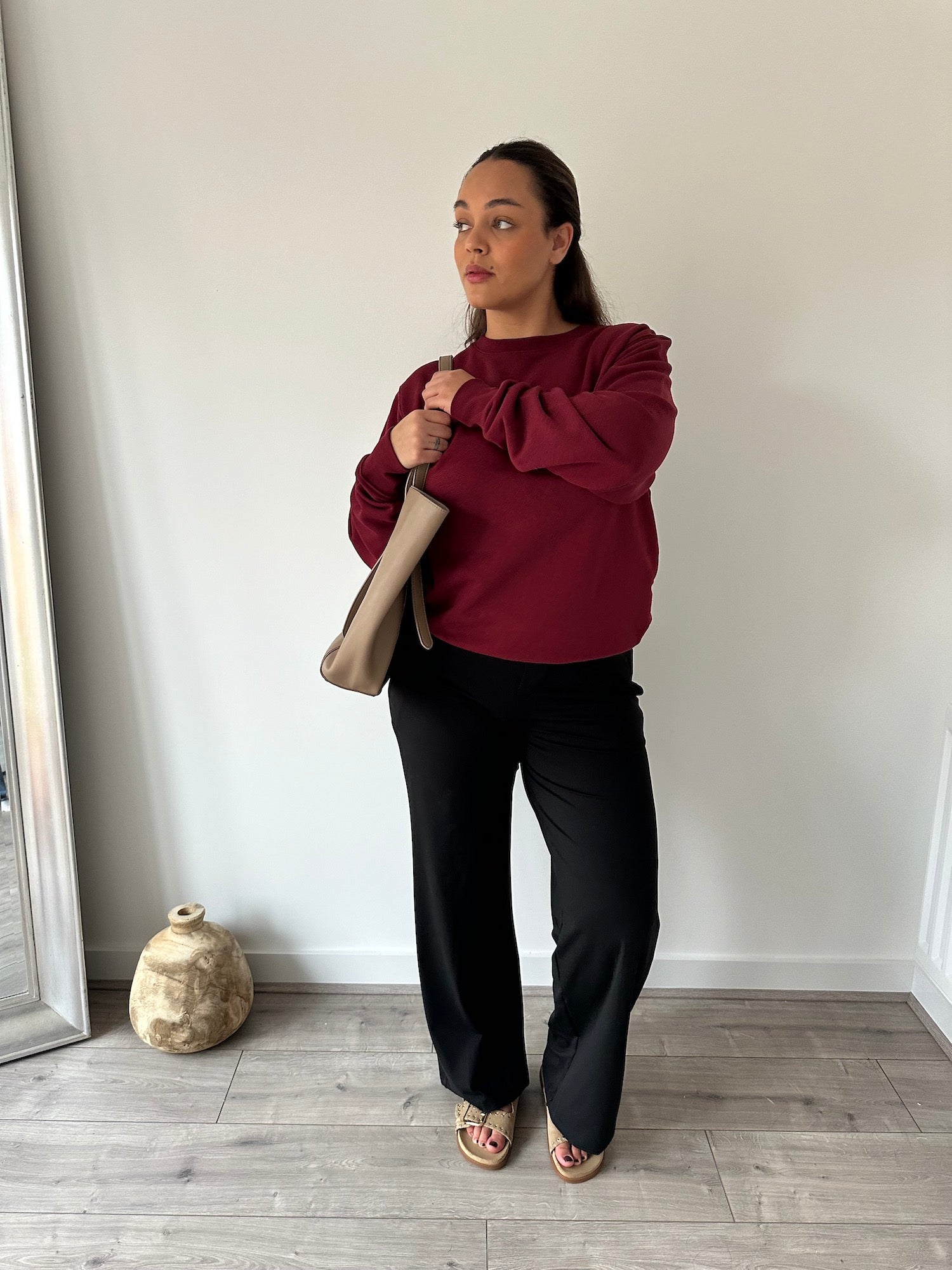 Eine Frau in einer schwarzen „Ninepine Asana Relaxed“-Hose, kombiniert mit einem burgunderroten Sweatshirt mit Rundhalsausschnitt und einer beigen Handtasche – ein schlichtes, minimalistisches Outfit für den Alltag