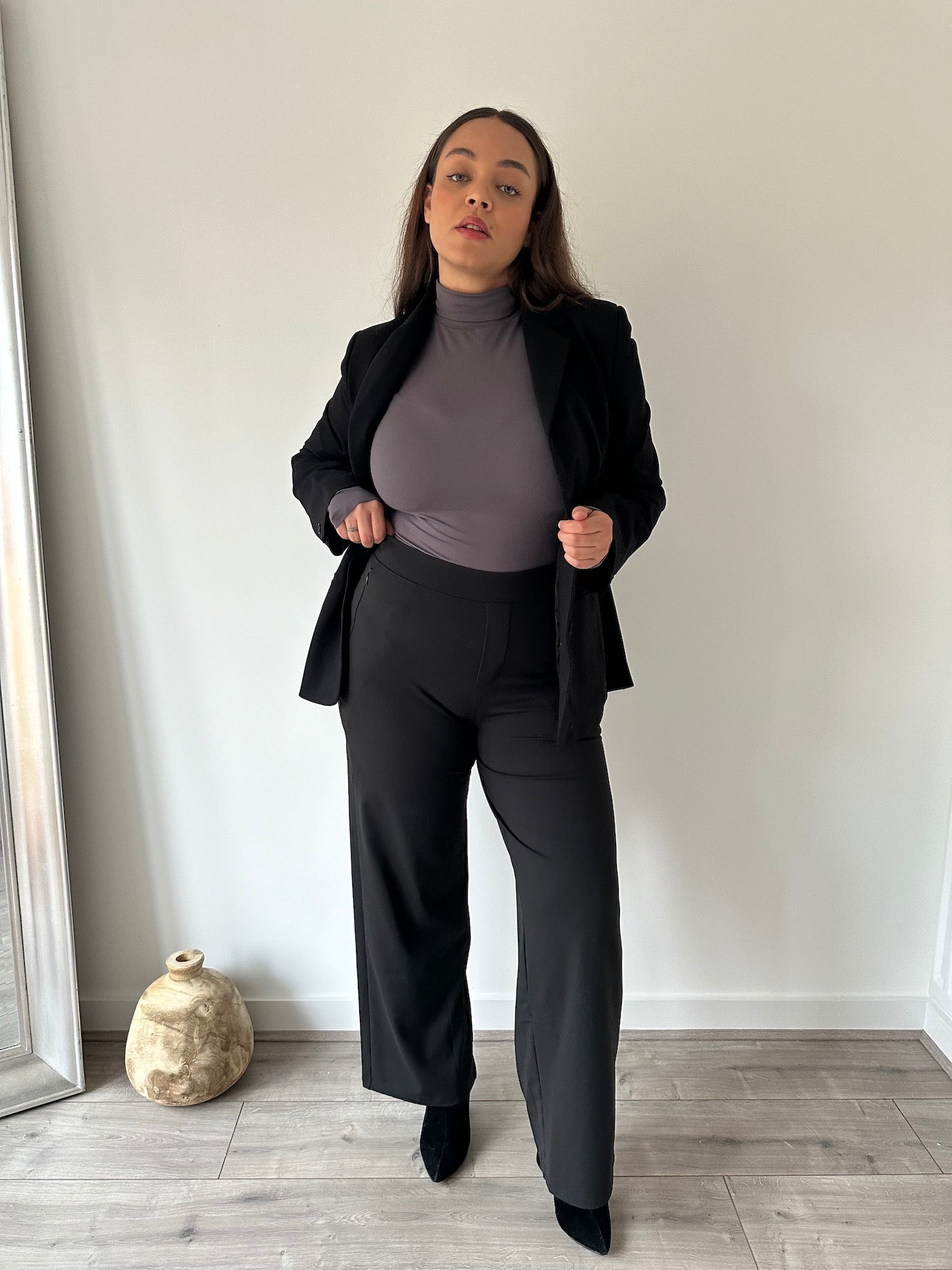 Eine kurvige Frau in einer schwarzen „Ninepine Asana Relaxed“-Hose, einem weichen Rollkragenpullover in Anthrazit und einem „Perfect“-Blazer präsentiert ein bequemes, maßgeschneidertes Outfit für den Alltag und das Büro