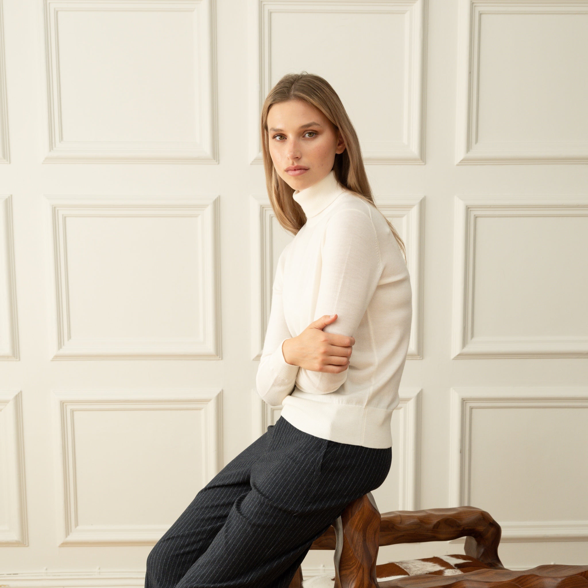 Woman wearing a Ninepine off-white merino wool relaxed turtleneck with dark pinstripe trousers against a wainscoted wall