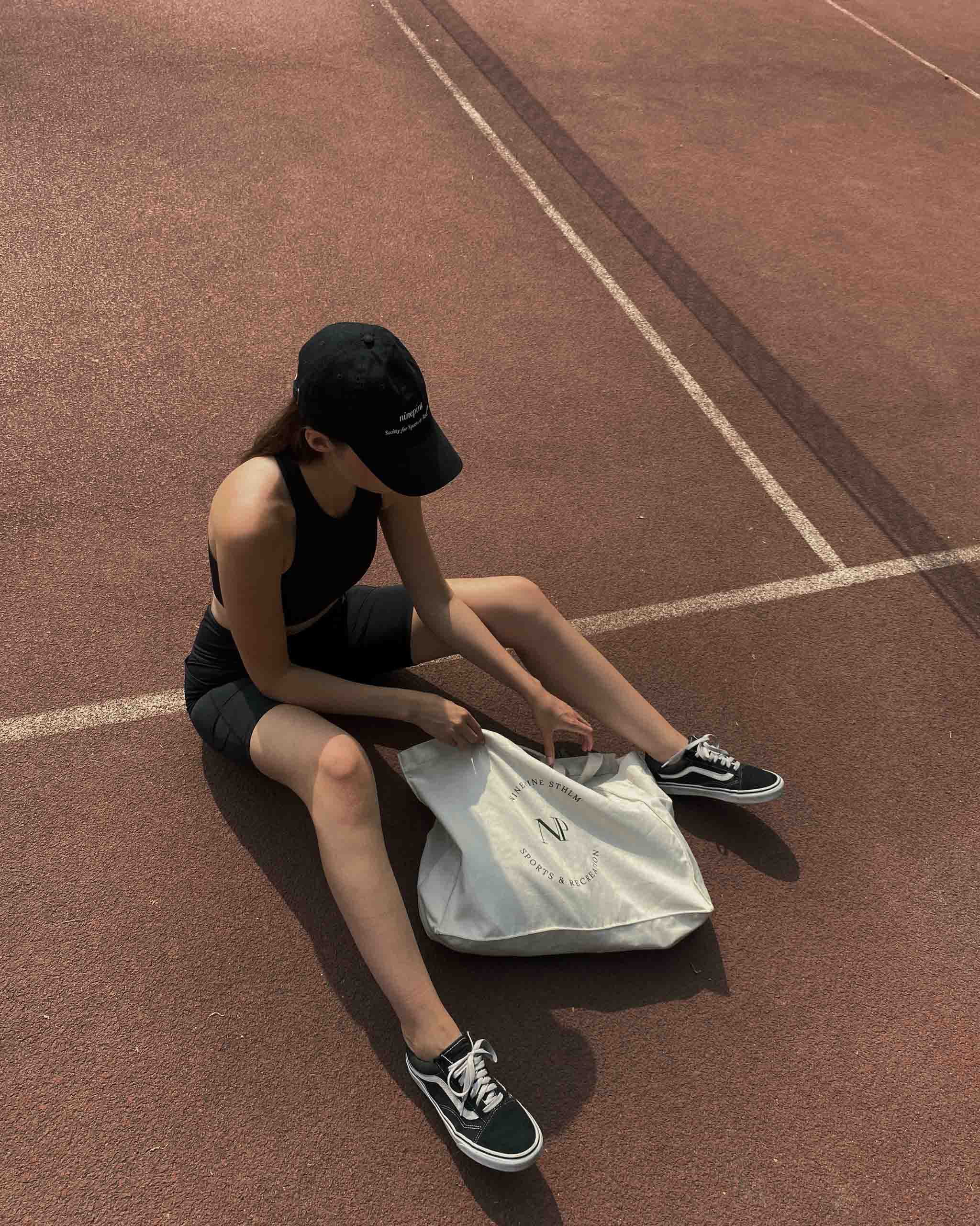 Girl in ninepine cap sitting on running track looking through her canvas tote bag