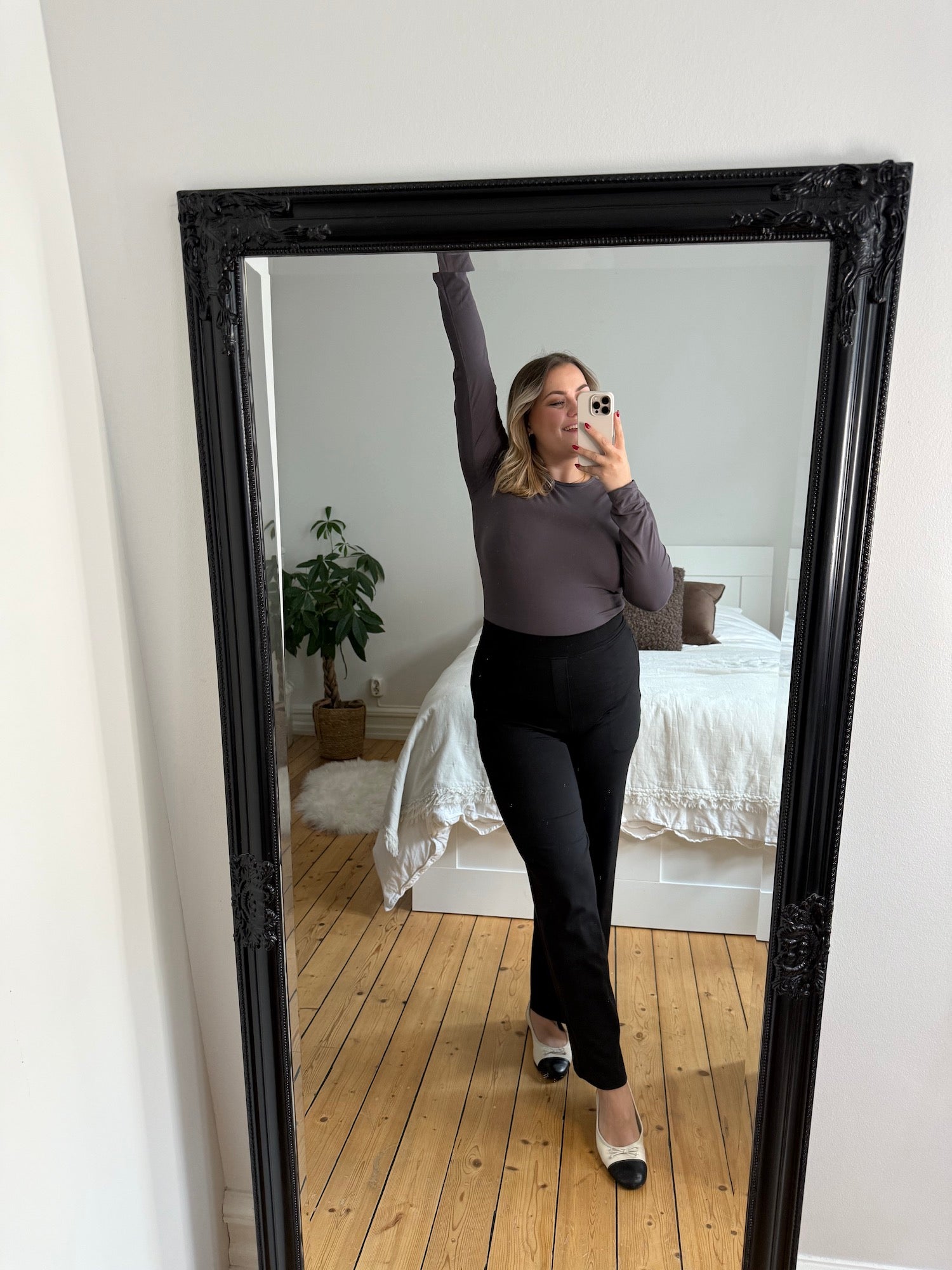 Eine Frau macht ein Spiegel-Selfie in einer schwarzen, taillierten, geraden Hose und einem eng anliegenden, langärmeligen Oberteil aus weichem Stoff, kombiniert mit zweifarbigen Ballerinas – ein schlichtes, minimalistisches Outfit für den Alltag