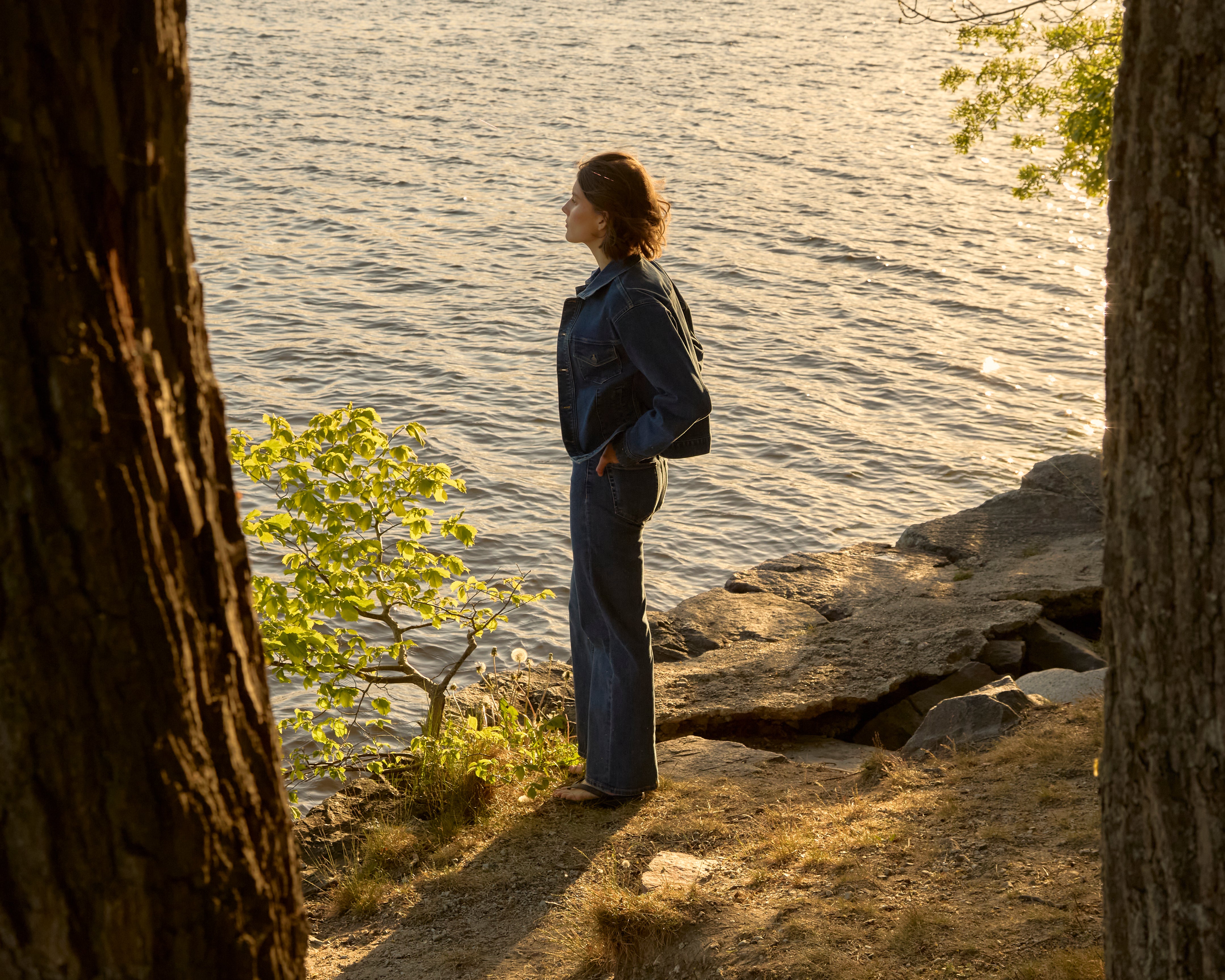 Eine Frau in einer mittelblauen Ninepine ComfortDenim-Jacke und einer lockeren Jeans steht in der goldenen Stunde an einem bewaldeten schwedischen Seeufer und blickt über das sonnenbeschienene Wasser.