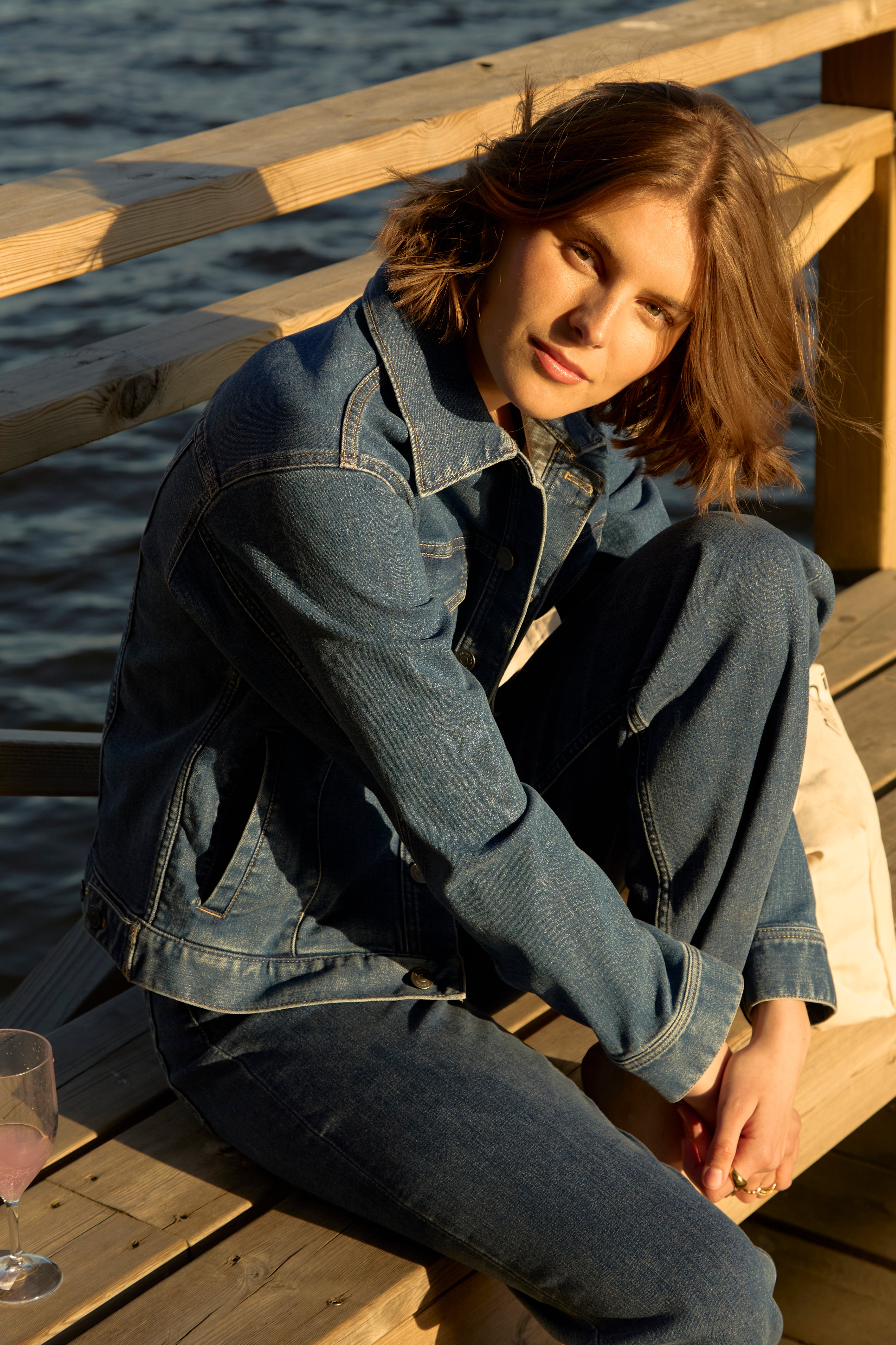 Frau sitzt in der goldenen Stunde auf einem hölzernen Pier an einem schwedischen See, gekleidet in eine mittelblaue Ninepine ComfortDenim-Jacke und dazu passende Jeans, entspannte Sommerabendstimmung.