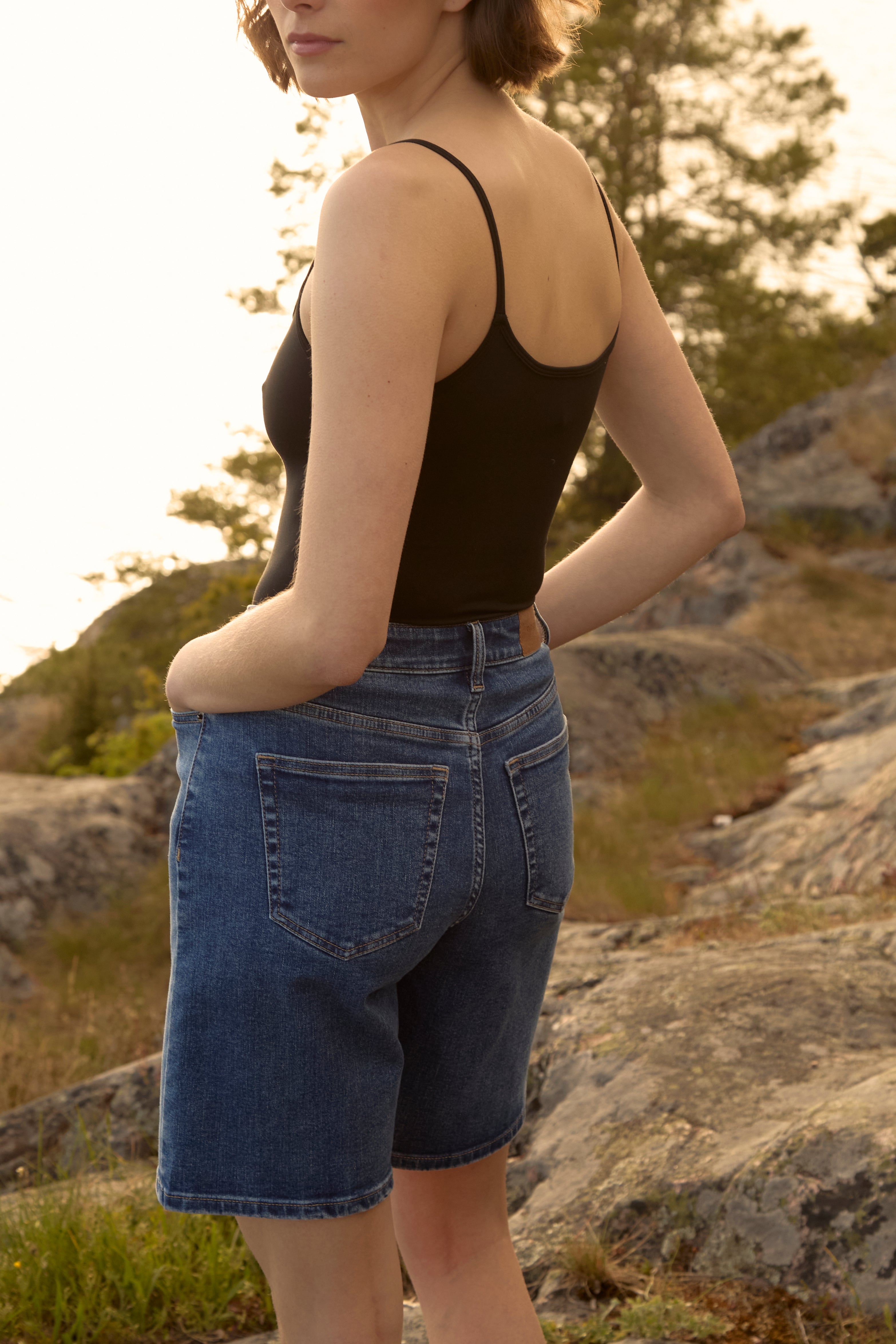 Frau bei goldenem Sonnenuntergang im schwedischen Archipel, bekleidet mit mittelblauen Ninepine ComfortDenim-Shorts und einem schwarzen Trägerhemd, entspannte Sommerstimmung.