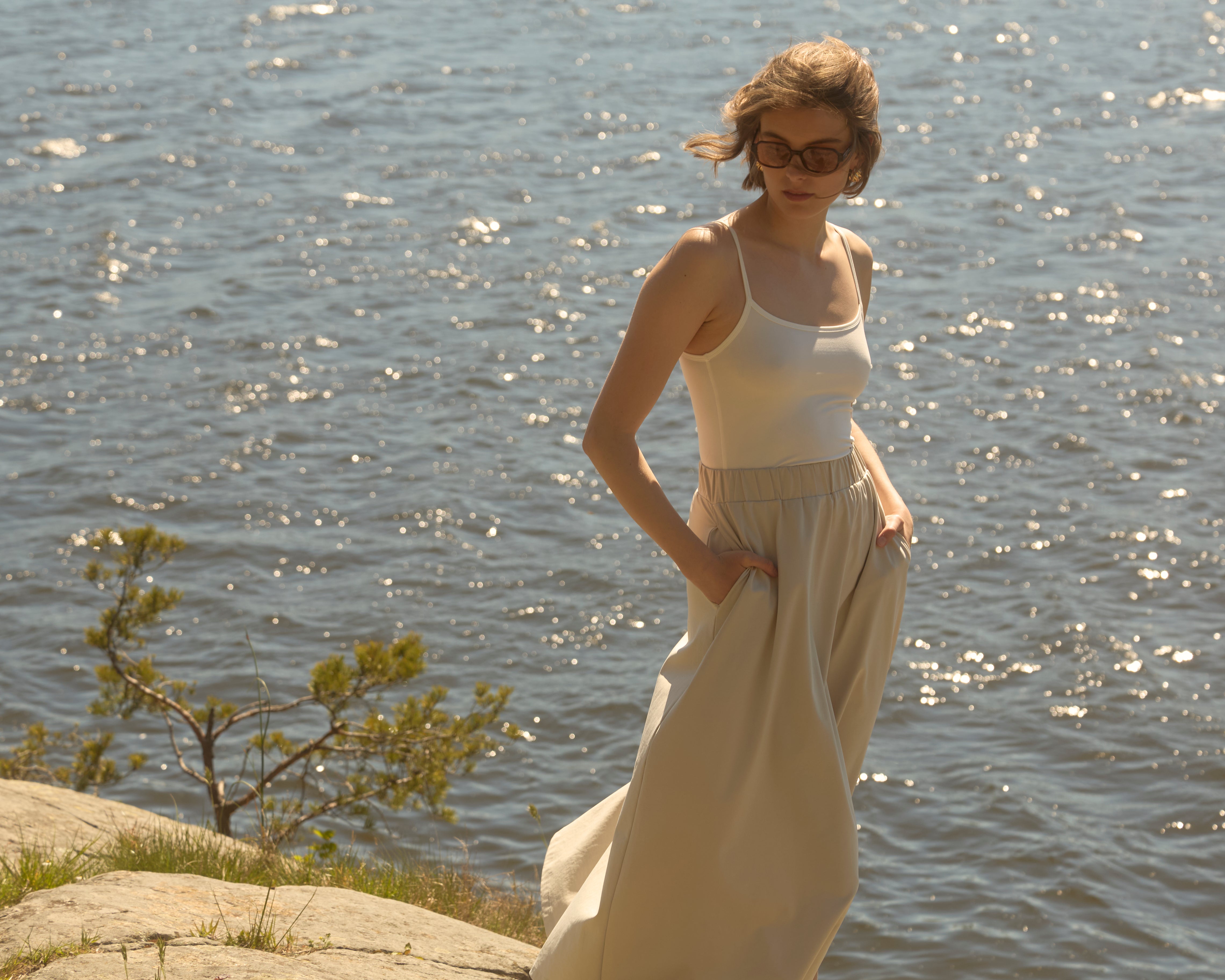 Eine vom Wind zerzauste Frau an einer glitzernden skandinavischen Küste, gekleidet in einen sandbeigen, fließenden Rock von Ninepine und ein weißes Tanktop – ein luftiger Sommerlook.
