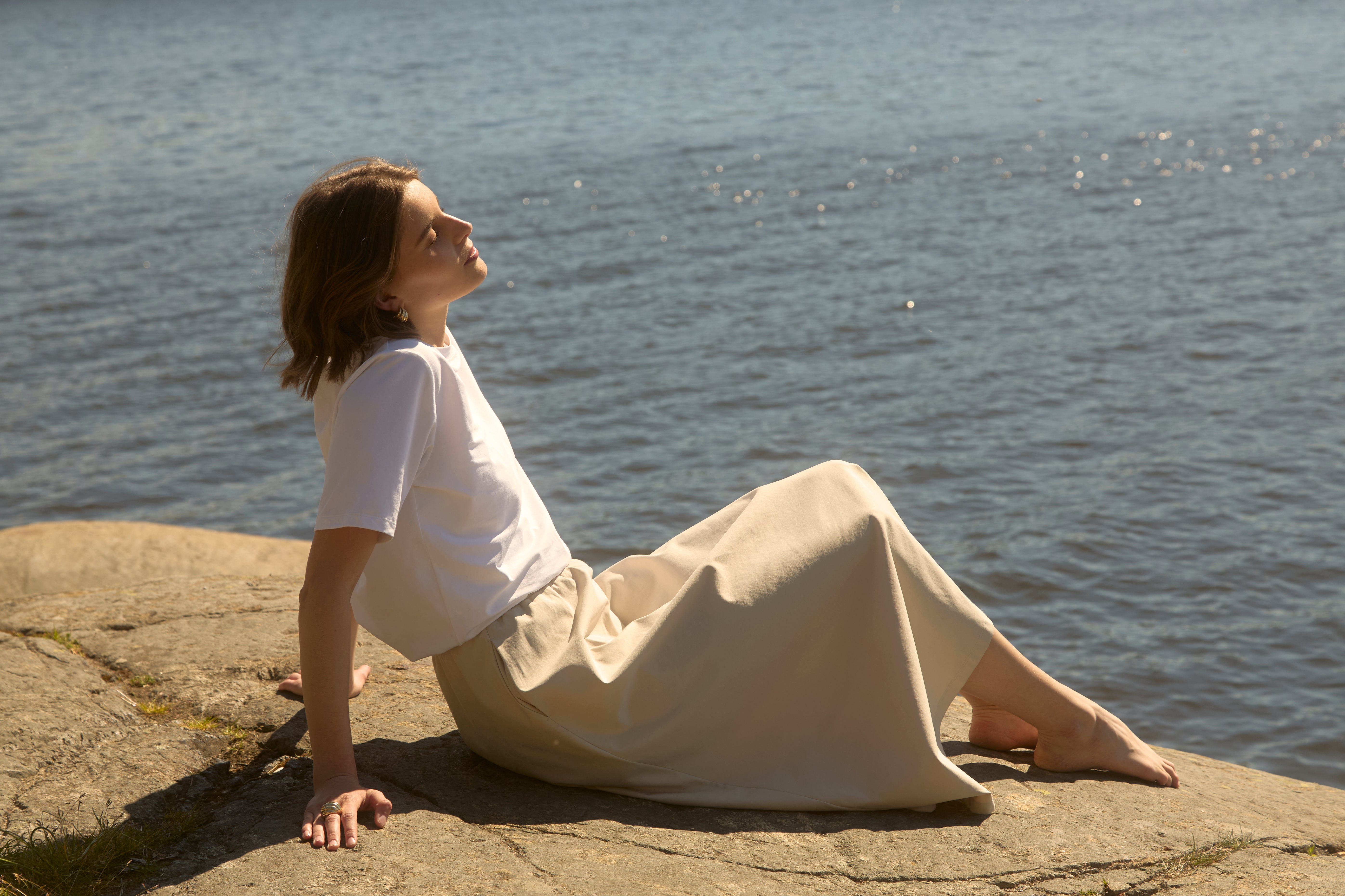 Frau in einem beigen, fließenden Ninepine Flowy-Rock und einem weißen T-Shirt, barfuß an einem sonnengewärmten nordischen Strand liegend, ruhiger Sommer-Minimalismus