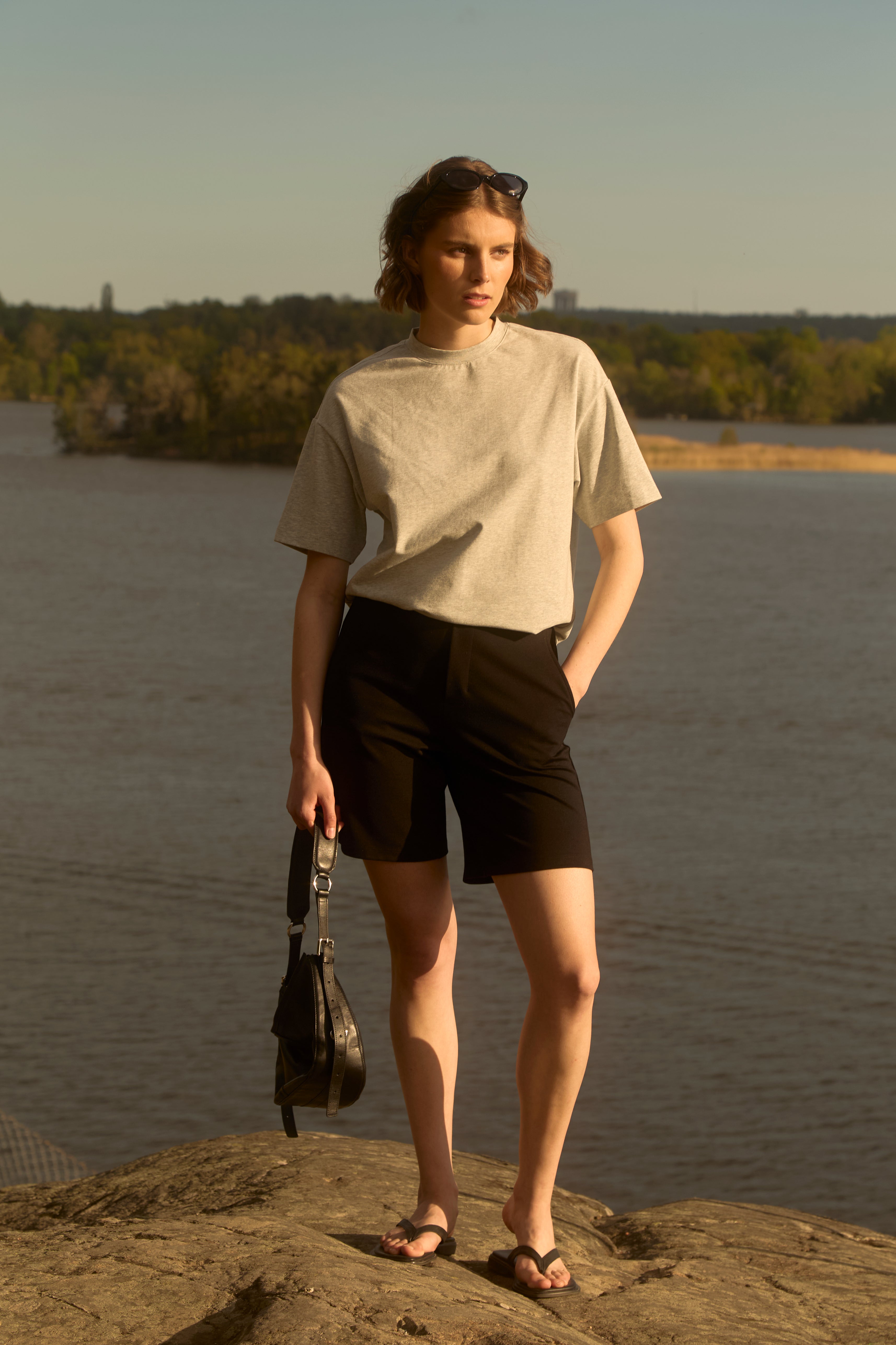 Frau am Ufer eines schwedischen Sees zur goldenen Stunde, bekleidet mit schwarzen Ninepine Light Ponte-Shorts und einem grauen Pima-T-Shirt, eine kleine Tasche in der Hand – ein klarer nordischer Sommerlook.