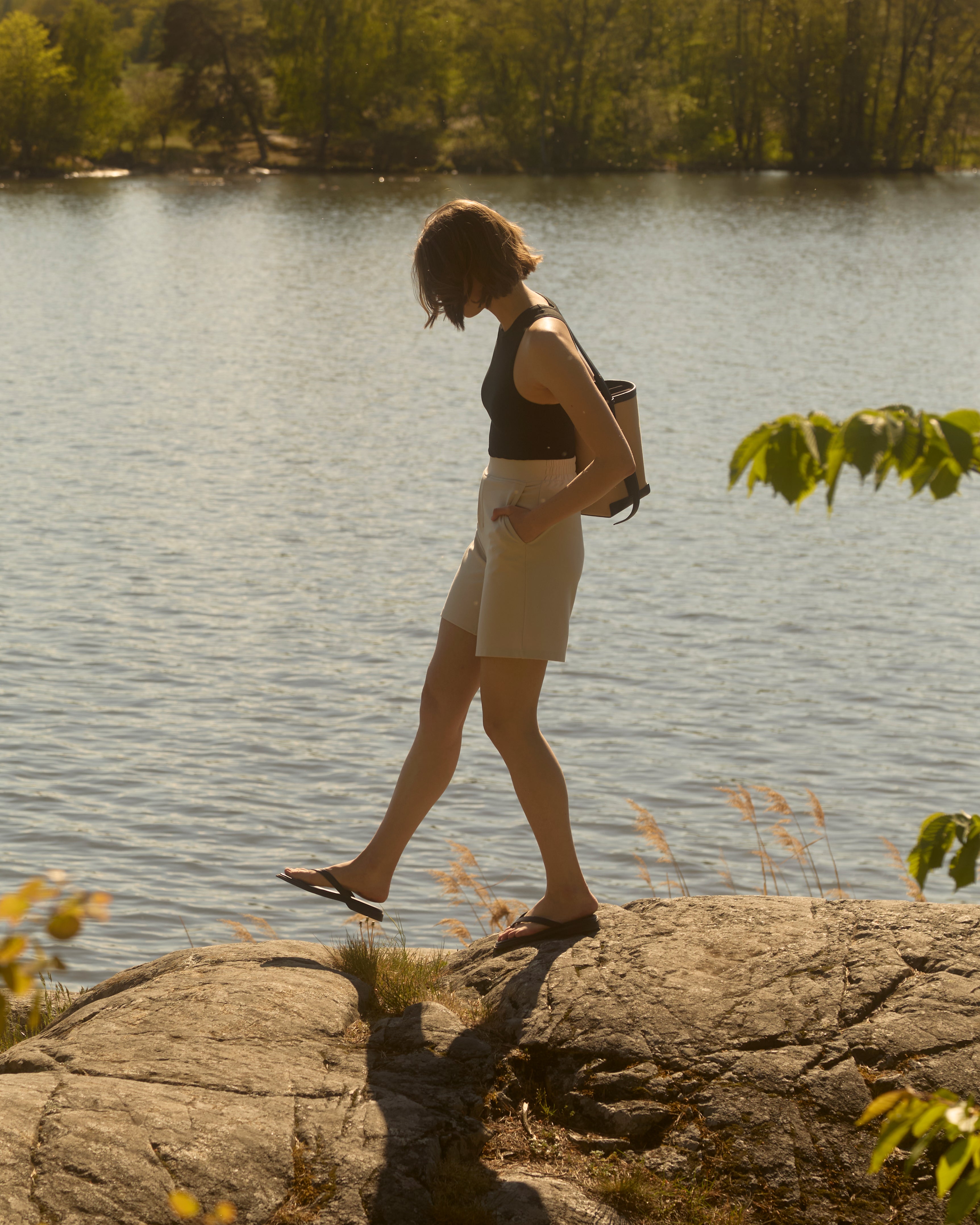 Seitenprofil einer Frau, die in sandbeigen Ninepine Light Ponte-Shorts, einem schwarzen Racerback-Tanktop und Flip-Flops an einem felsigen Seeufer entlangläuft – entspannte schwedische Sommerstimmung.