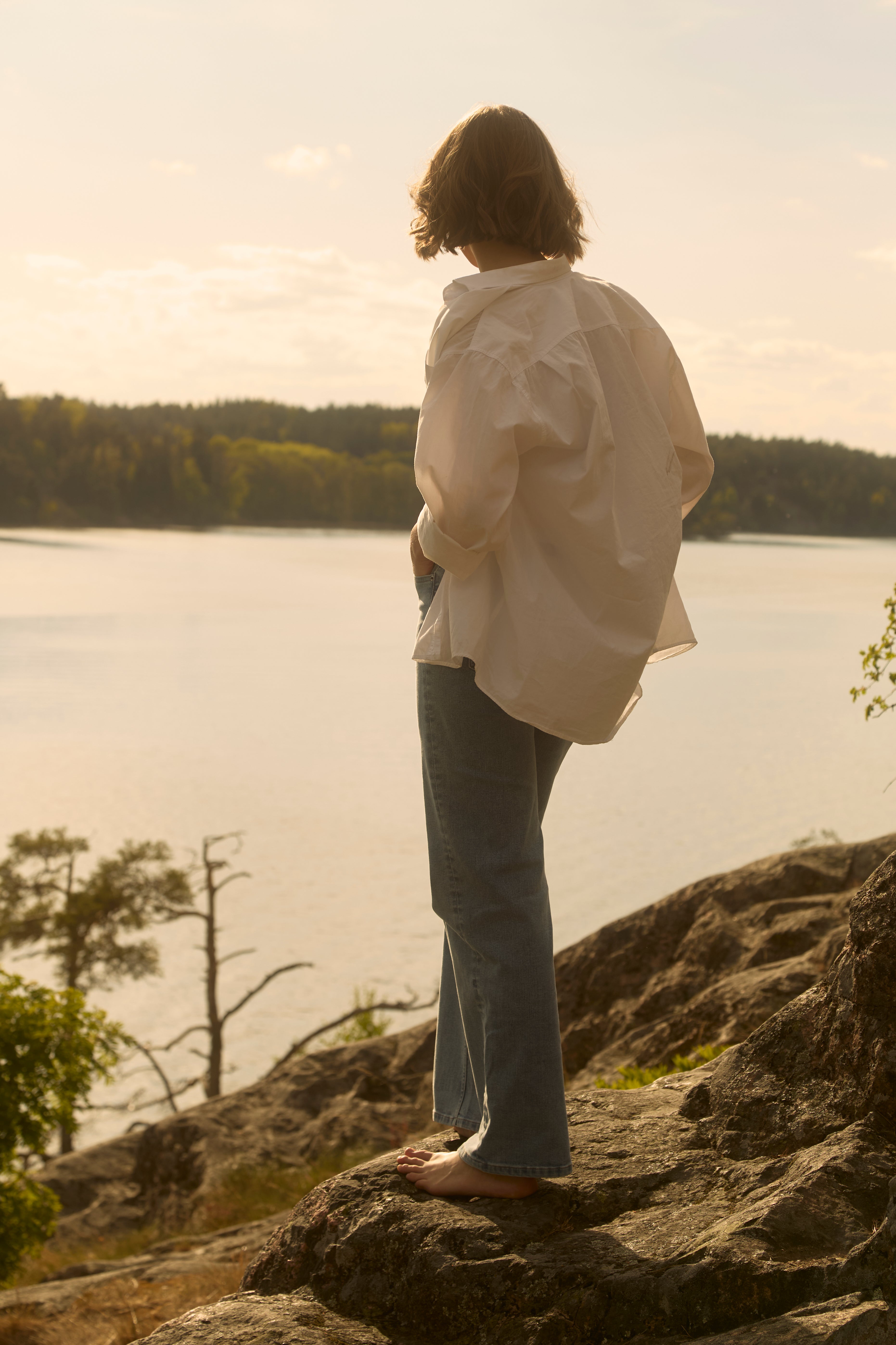Eine barfüßige Frau in hellblauen Ninepine Relaxed ComfortDenim-Jeans und einem weiten weißen Hemd steht an einem felsigen schwedischen Seeufer zur goldenen Stunde des Hochsommers.
