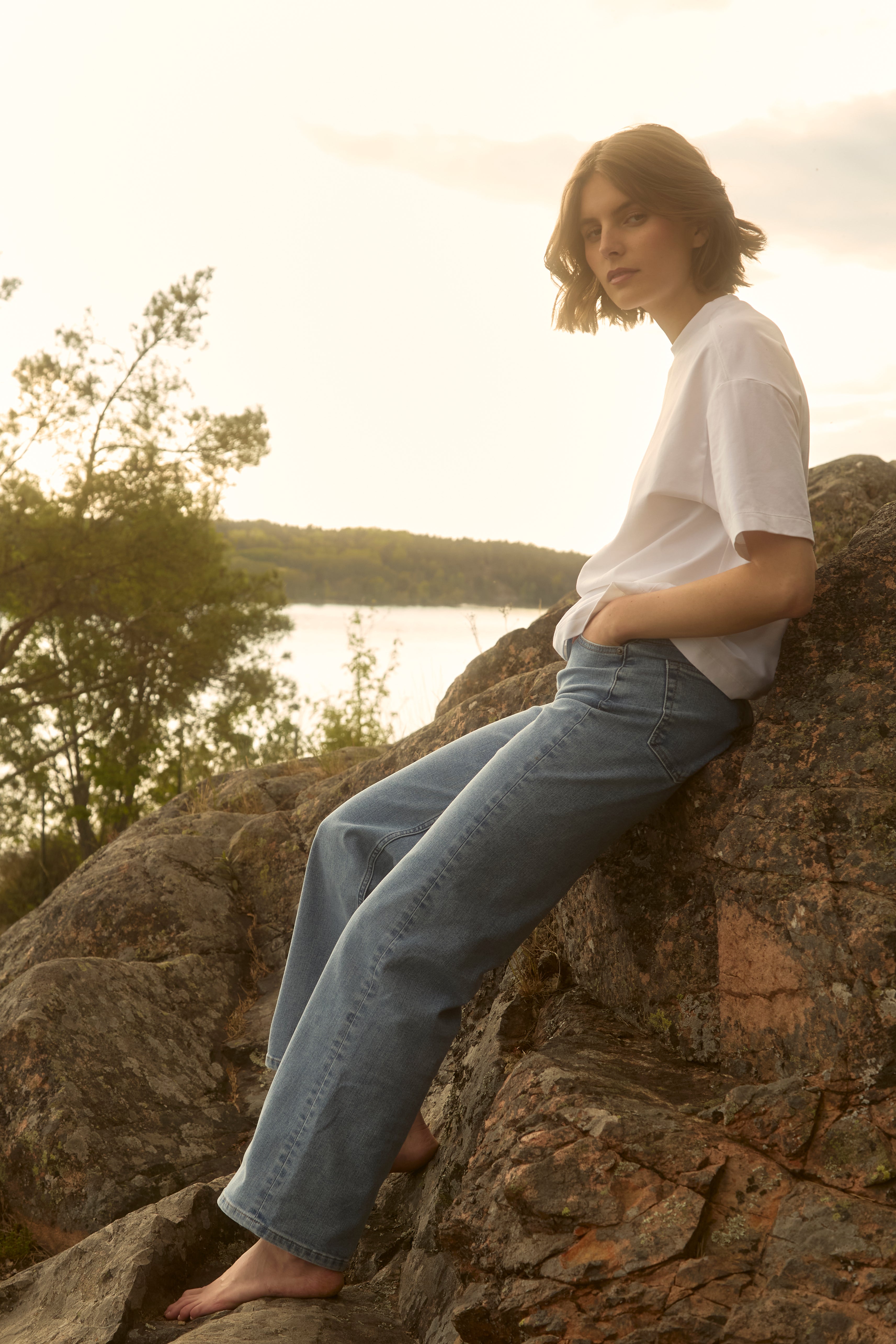 Eine barfüßige Frau in hellblauen Ninepine Relaxed ComfortDenim-Jeans und einem knackig weißen Pima-T-Shirt liegt bei Sonnenuntergang auf einem sonnengewärmten Felsen am Ufer eines schwedischen Sees und genießt die ruhige Sommerstimmung.